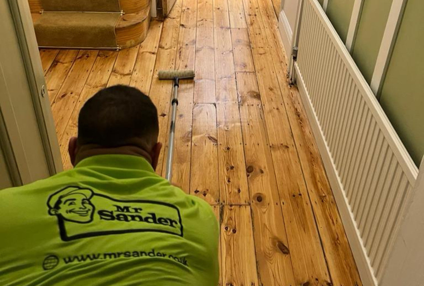 Professional man from Mr Sander® wearing a lime green shirt with the Mr Sander® logo, waxing a pine floor with a roller in an Arkley, UK home. The image shows meticulous application of lacquer to achieve a smooth and protective finish on the wooden floor.