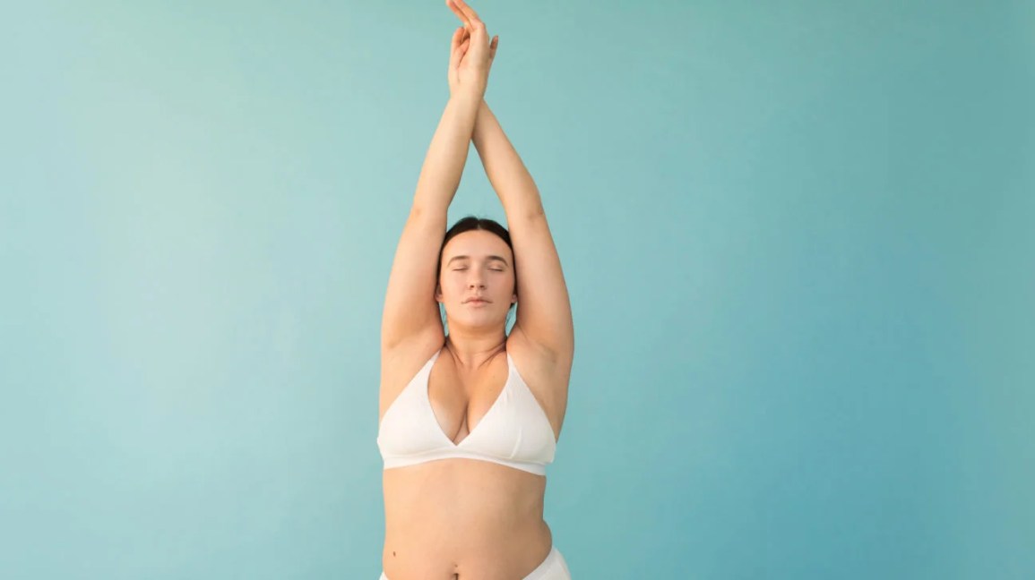 plus-size model wearing white undergarments with arms raised overhead against a light blue background