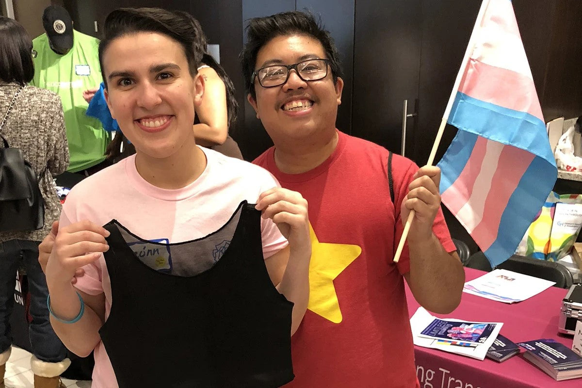 Smiling trans folks hold up a free chest binder and trans pride flag. (Courtesy of Point of Pride.)