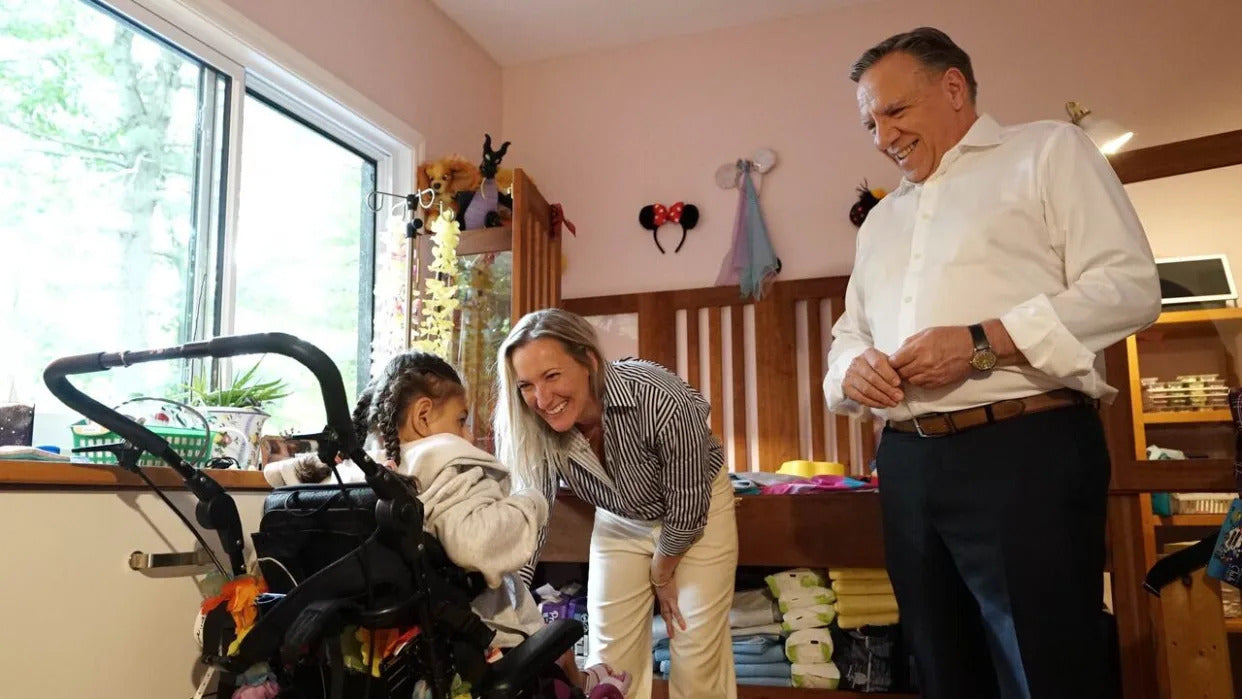 Soulanges MNA Marilyne Picard stands before her daughter Dylane, who has a congenital disease which prevents her from walking and speaking, as Premier François Legault looks on. (Mathieu Potvin/Radio-Canada - image credit)