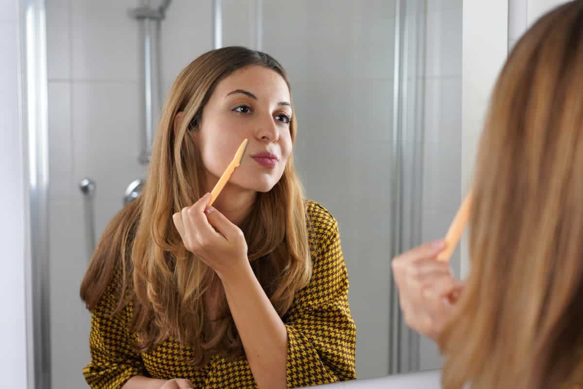 Beautiful young woman using razor while looking in the mirror on the bathroom