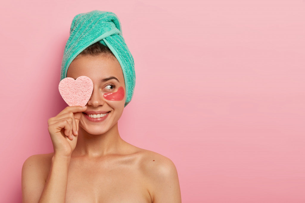 smiling woman holding heart-shaped pink sponge in face with towel in hair